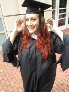 Photo of a white woman with long red hair in a graduation cap and gown, smiling with her hands up as if she was adjusting the cap